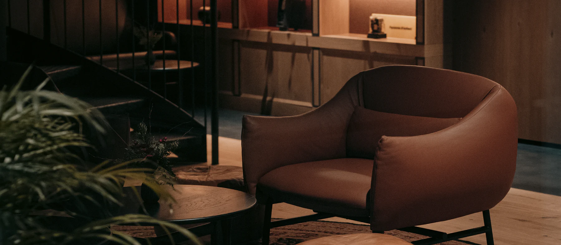 Un sillón de piel bajo luces cálidas, con plantas y estanterías de madera al fondo.
